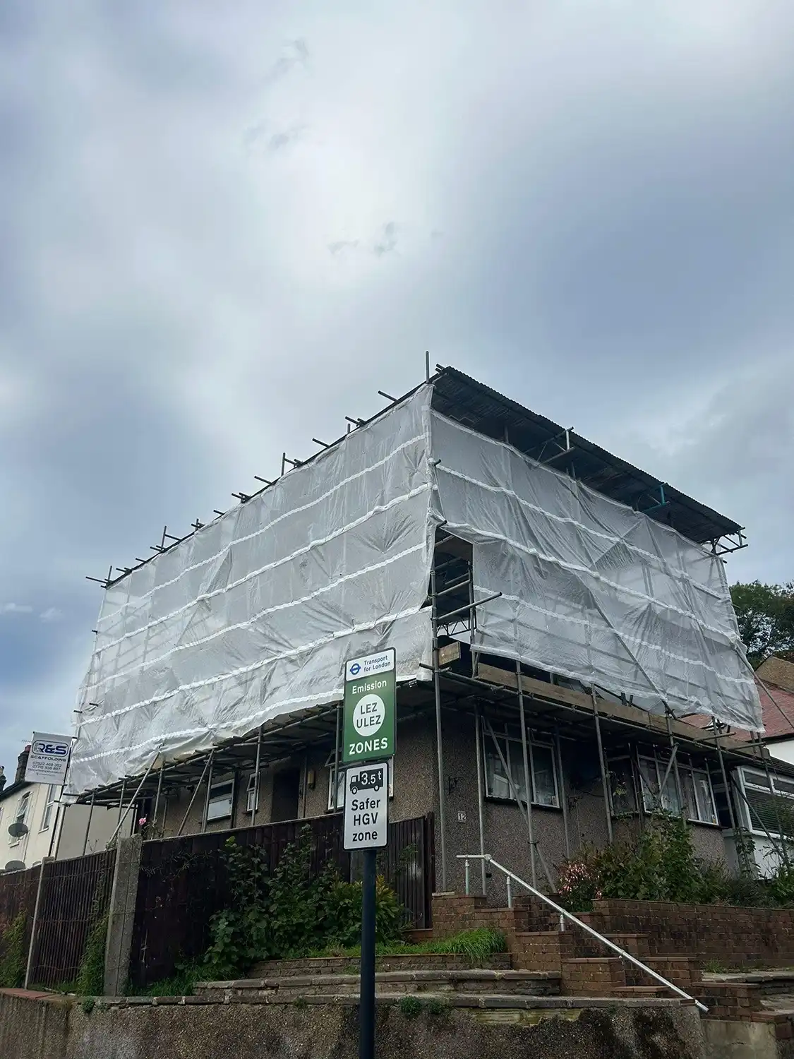 House covered with scaffolding and white protective sheeting under a cloudy sky.