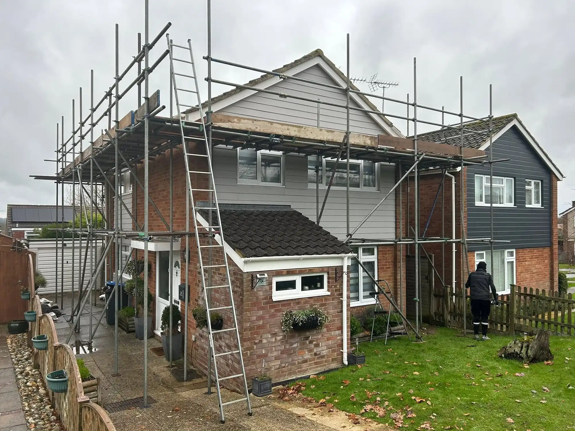 Two-story brick and siding house with scaffolding and a person opening a wooden gate in the yard.