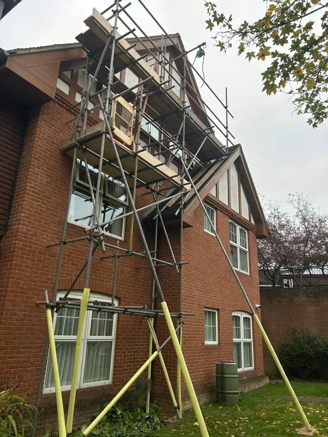 Scaffolding erected against a brick house with multiple windows and a green water barrel outside.