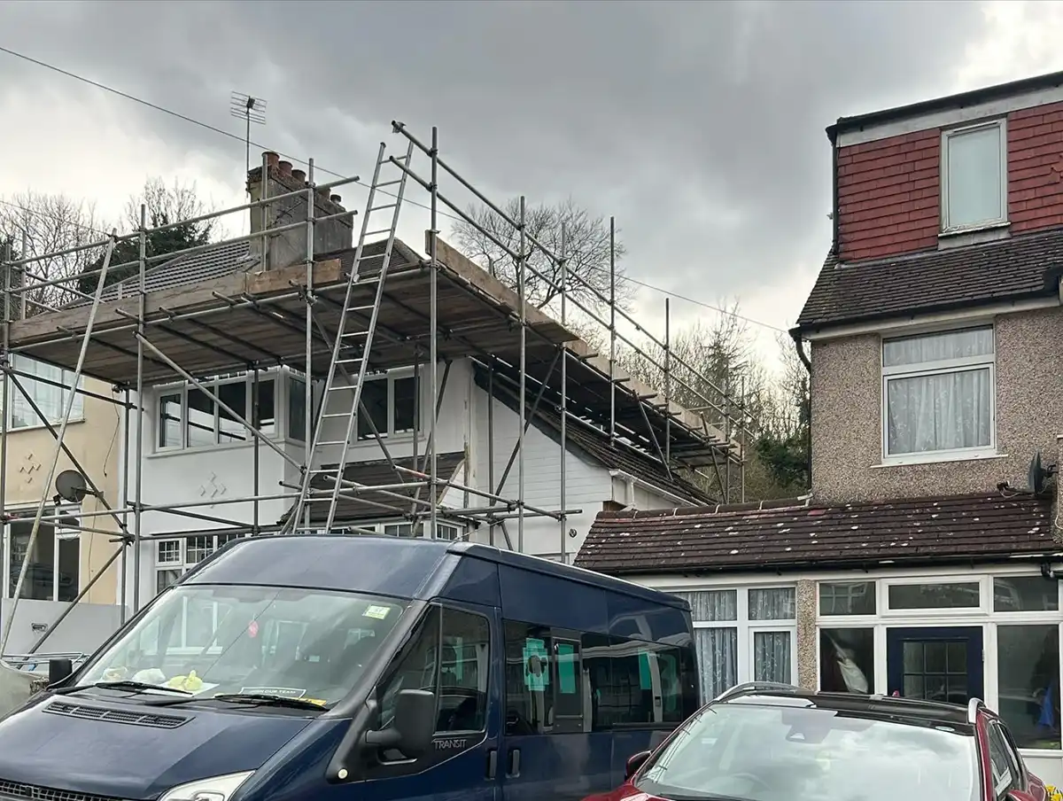 Two houses with scaffolding on one under cloudy sky and parked blue van and red car.