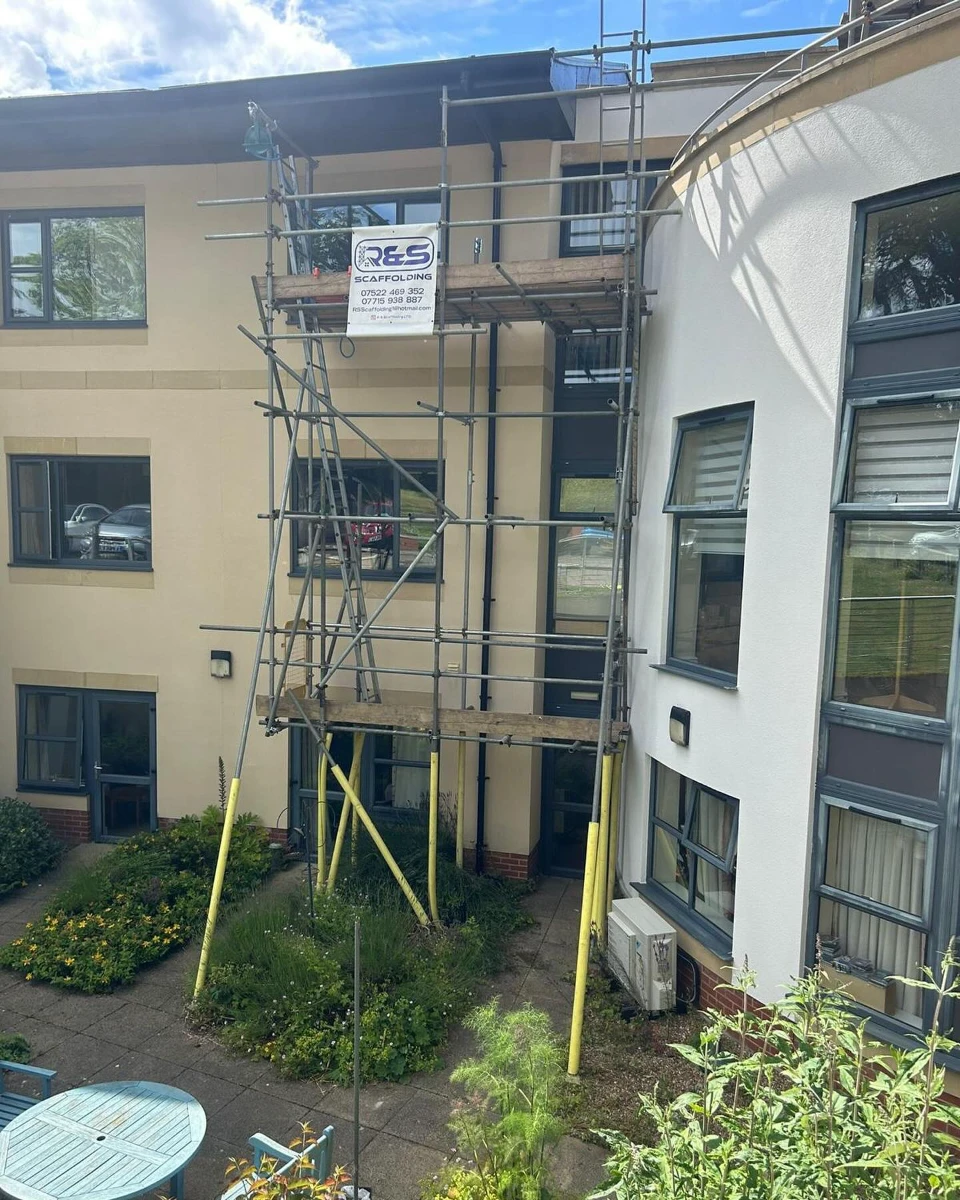 Scaffolding with R&S Scaffolding sign set up outside a beige multi-story building with garden and patio.