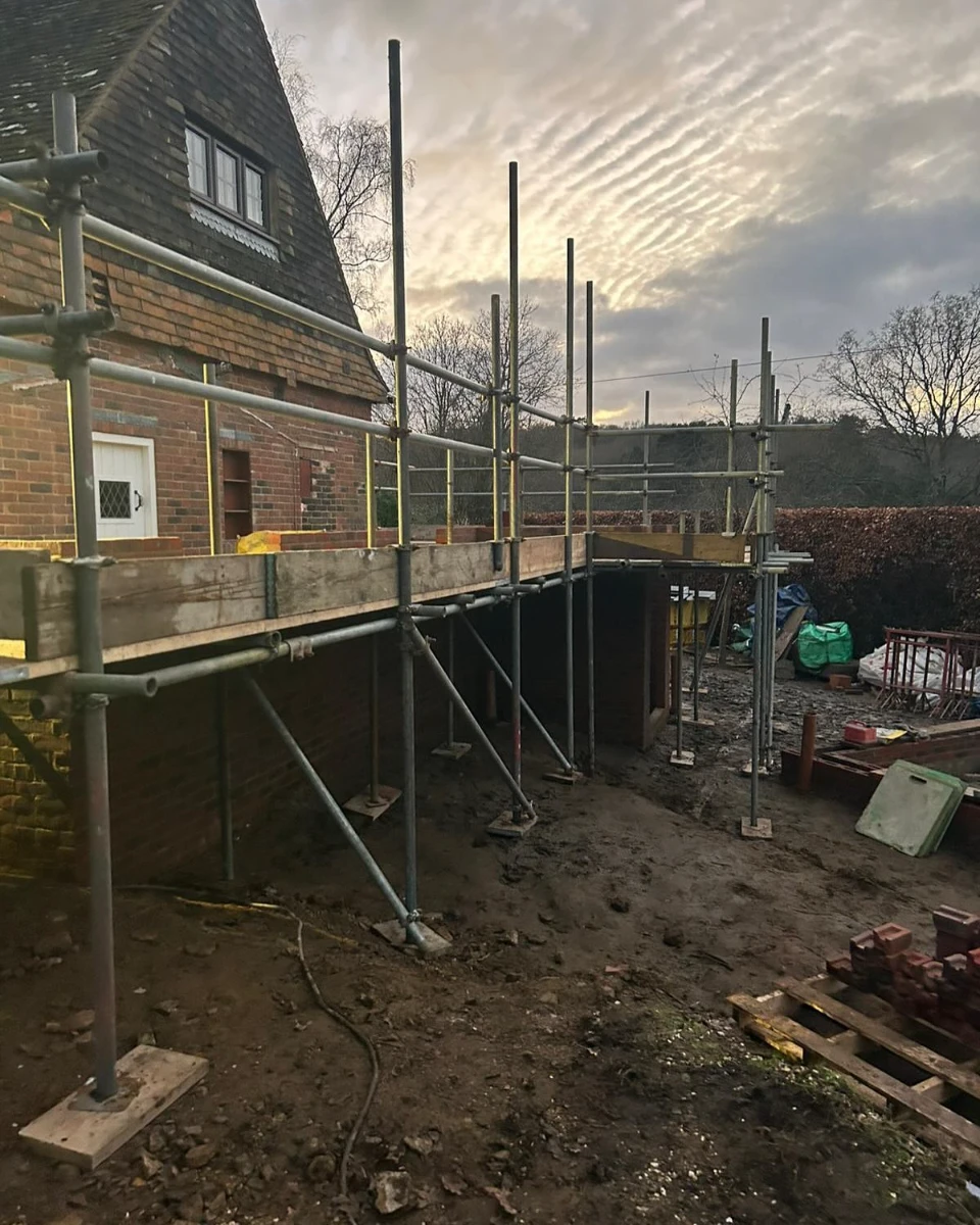 Scaffolding erected along a brick house with bare ground and construction materials nearby at dusk.