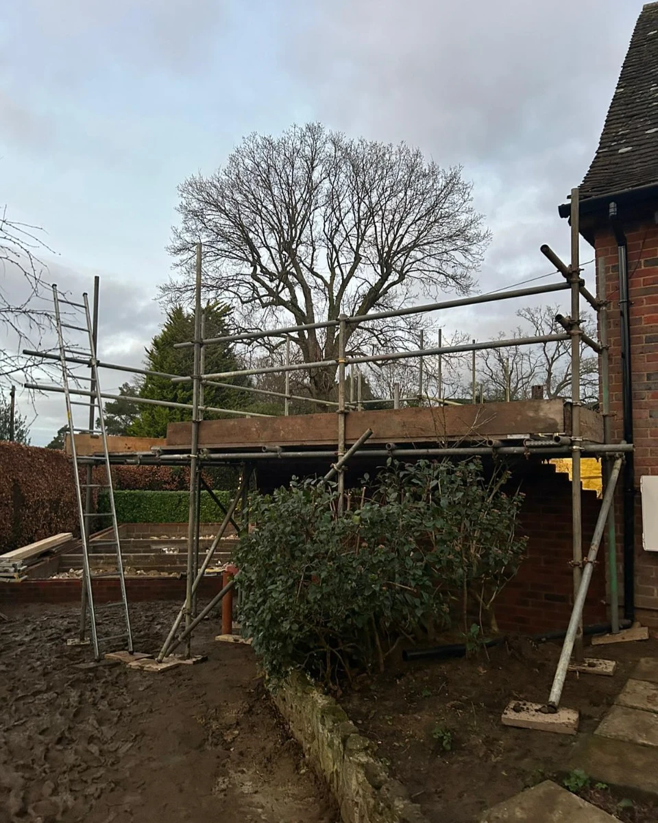 Scaffolding set up around a brick extension under construction beside a house with bare trees in background.
