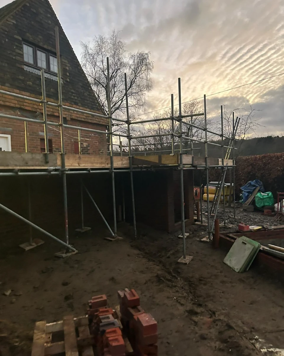 Scaffolding assembled around a brick building with a muddy construction site in front at dusk.
