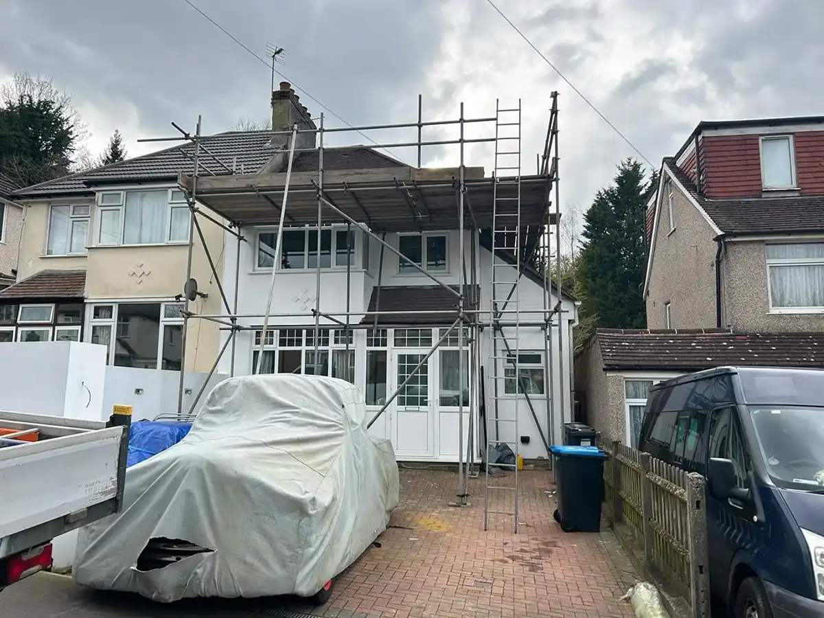 House with scaffolding on the front during overcast weather; covered car and parked van nearby.