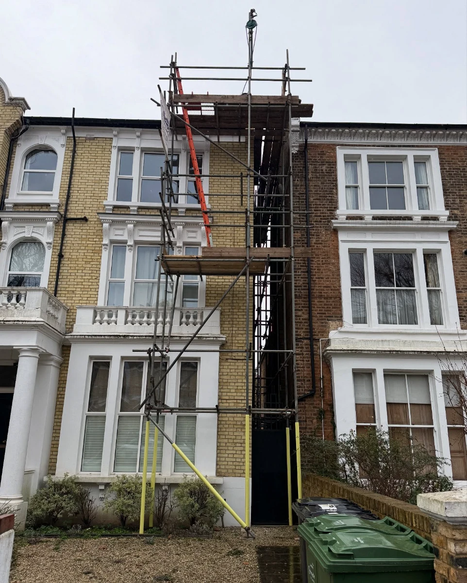  Scaffold structure with a red ladder between two multi-story brick townhouses on a cloudy day.