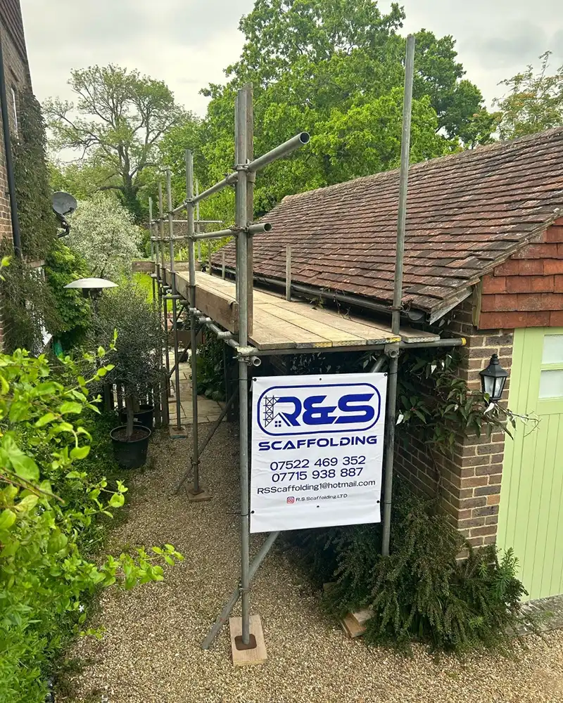 Metal scaffolding set up along a narrow gravel path beside a brick building with a pitched roof.