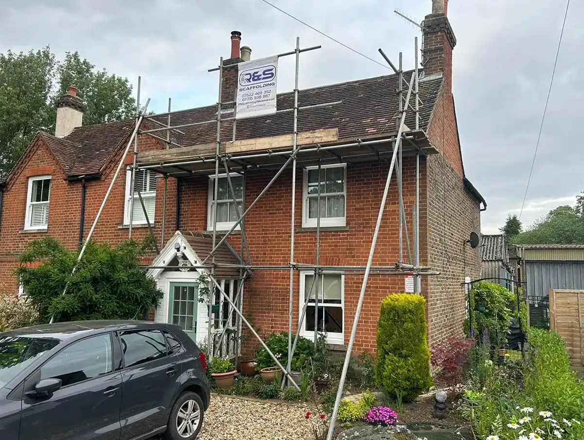 Red brick house with scaffolding on the front wall and a black car parked outside on gravel.